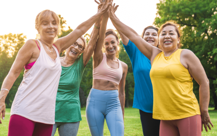 A group of five women in athletic clothing joyfully joining hands in a circle during a sunny outdoor yoga class.