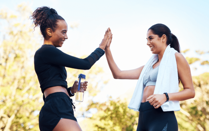 Two women in athletic gear are high-fiving each other outdoors, with a bottle of water in one hand and a towel draped around the other.